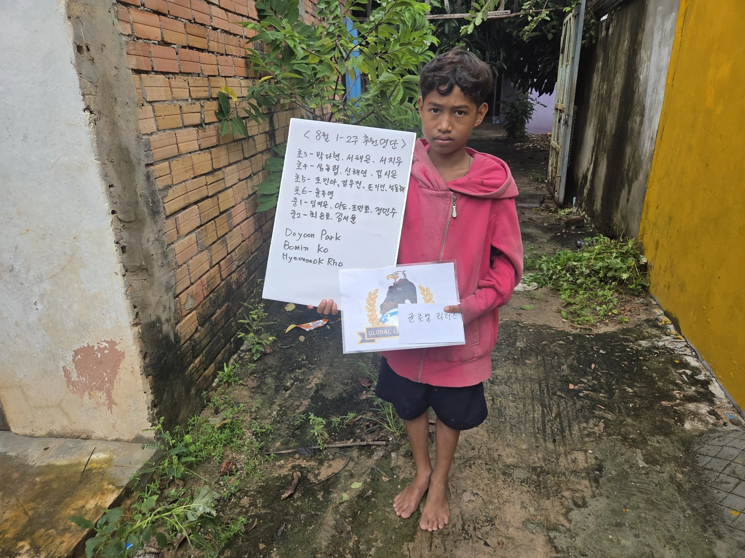 A young boy standing barefoot outdoors, holding a handwritten sign with Korean and English text, and a printed emblem featuring an eagle with a branch. The background shows a brick and concrete wall, green plants, and a yellow wall.
