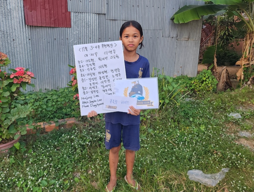 A young girl wearing a blue outfit standing in a garden, holding a sign with handwritten Korean text and an envelope with a decoration. Behind her are plants, flowers, a metal wall, and tropical foliage.
