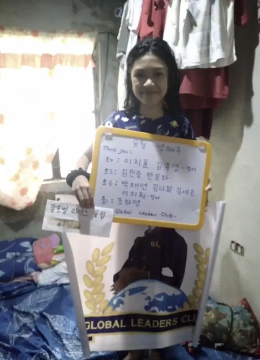 A girl holding a whiteboard and a paper in a room with hanging clothes and a bed. The whiteboard has writing in Korean. The girl is smiling, and there is a banner with the text "Global Leaders Club" and an image of a person with hands on a globe.