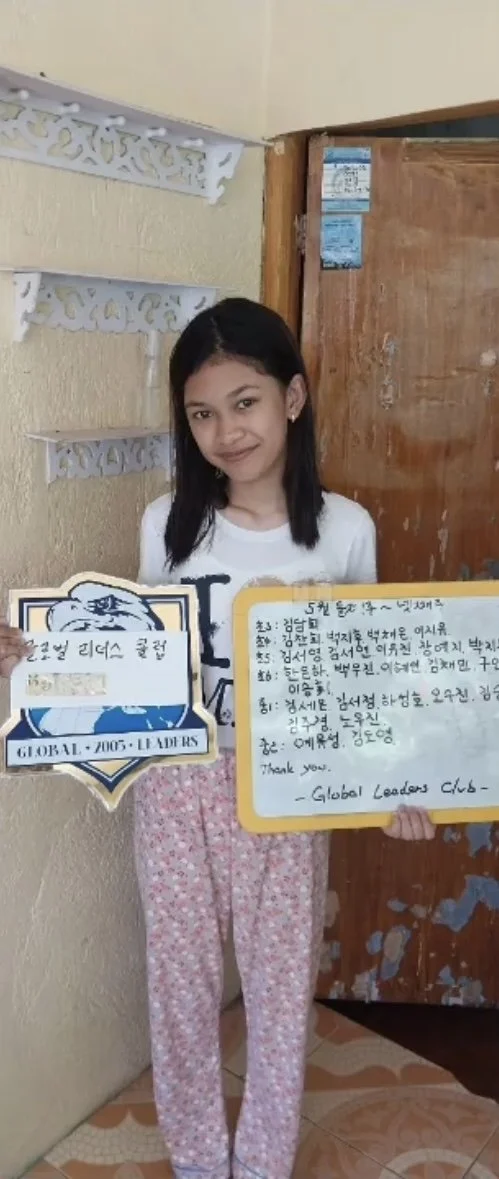 A young girl with black hair standing indoors against a beige wall, holding a certificate or plaque and a large handwritten note or letter in Korean. She is wearing pink pajama pants and a white shirt, smiling slightly.