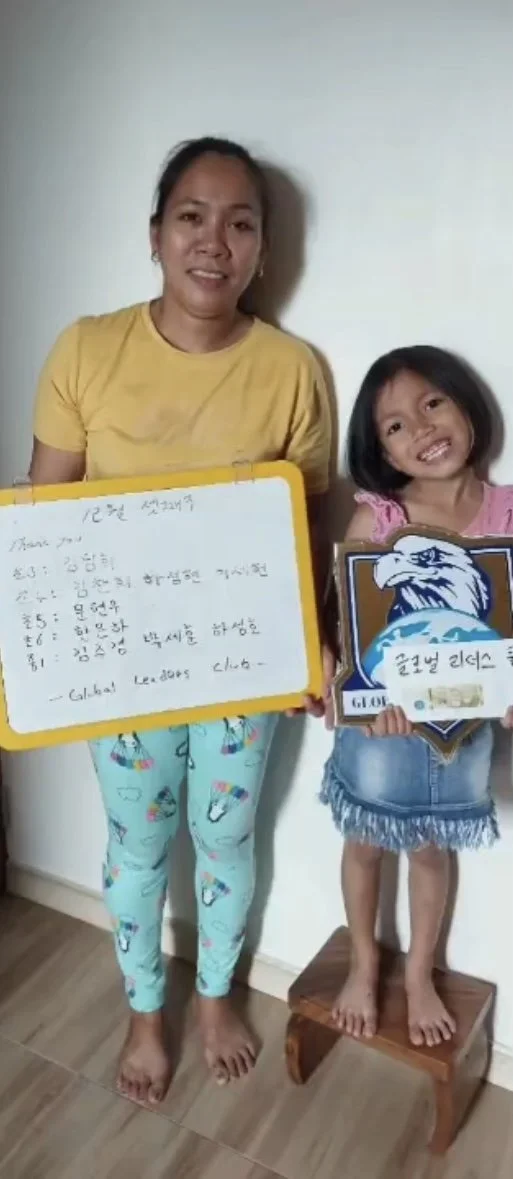 A woman and a young girl standing together indoors. The woman holds a whiteboard with handwritten notes, while the girl holds a plaque with an eagle graphic, standing on a small wooden stool. Both are smiling.