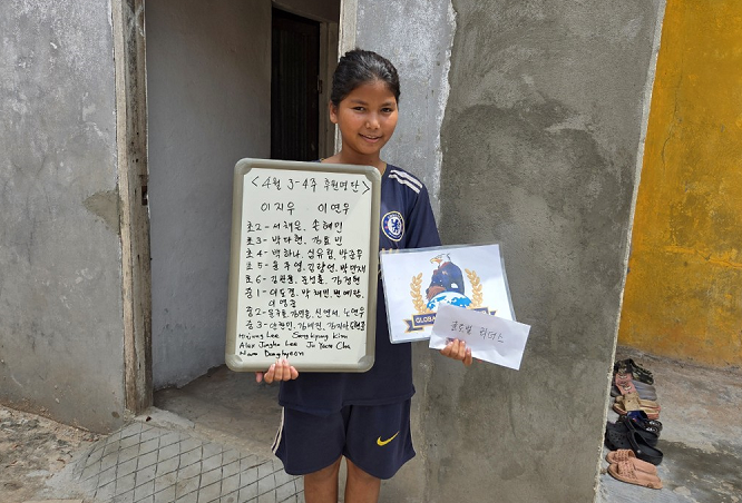 A young girl in a dark sports uniform smiling and holding a whiteboard with writing on it and a sign with a whale logo, standing in front of a rough concrete wall with some shoes on the ground nearby.