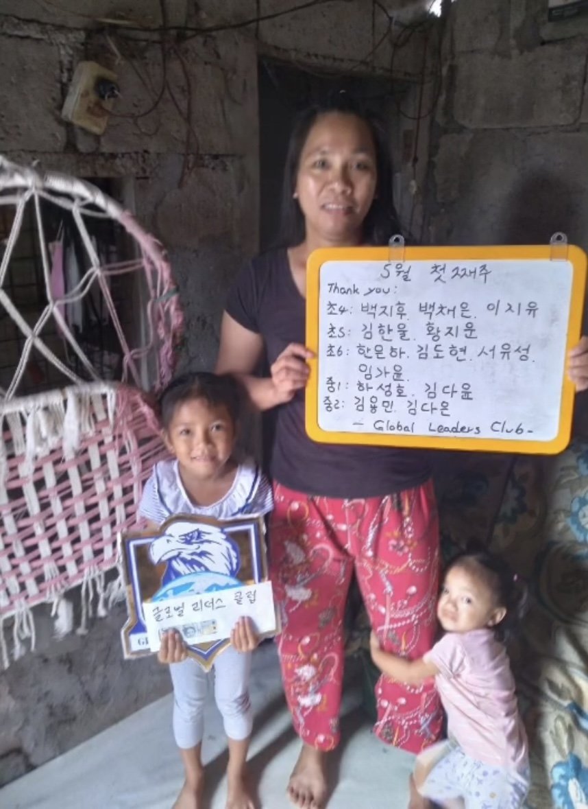 A woman and two young girls in a rustic room with stone walls. The woman is holding a whiteboard with handwritten notes and a message. The girl on the left is holding a sign with an eagle and Korean text, and the girl on the right is hugging the woma