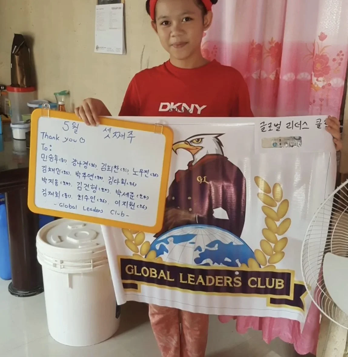 A young boy wearing a red DKNY shirt and a red headband, holding a large banner with the Global Leaders Club logo. The banner features an eagle illustration and a globe, and a handwritten note with Korean text and a thank you message.