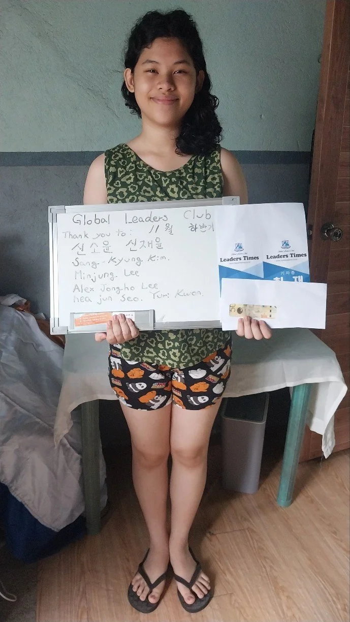 A young girl standing indoors, holding a whiteboard with handwritten notes and some blue and white paper. She is smiling, wearing a green sleeveless top with a leopard print pattern, black shorts with orange and white patches, and black flip-flops. T
