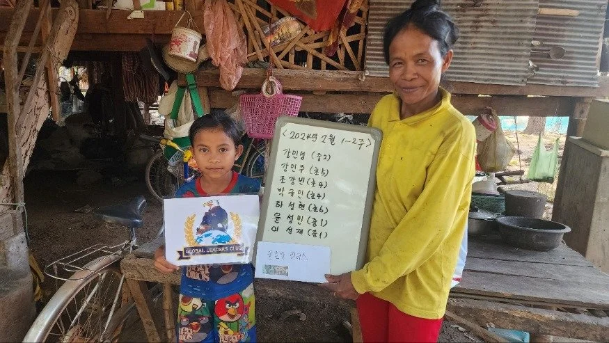 A young boy and an older woman standing inside a rustic wooden structure. The boy is holding a certificate and the woman is holding a whiteboard with writing on it. Various household items are visible in the background.