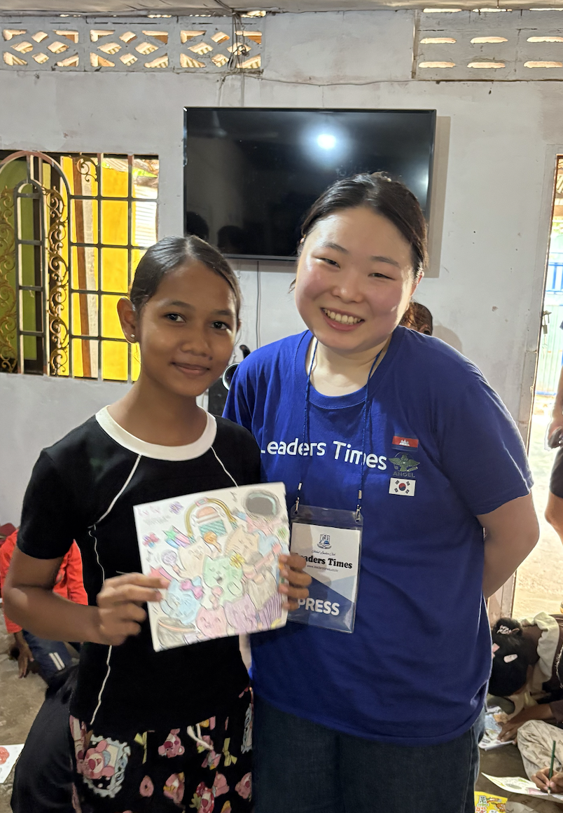 A young girl and a smiling woman standing indoors, with the girl holding a colorful drawing or card. The woman is wearing a blue t-shirt with 