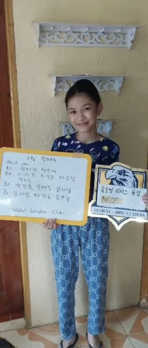 A young girl in blue pajamas holding a handwritten note and a sports team pennant, standing indoors against a yellow wall with decorative wall shelves above her.