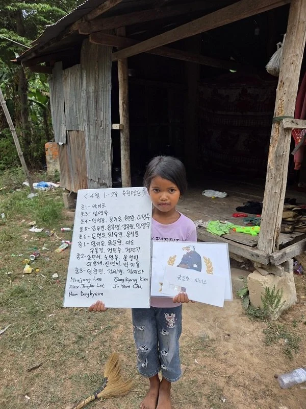 A young girl standing outside a wooden hut, holding handwritten notes on white paper. The girl has dark hair, is wearing a purple shirt and ripped jeans, and is barefoot. The background shows trees and scattered trash on the ground.