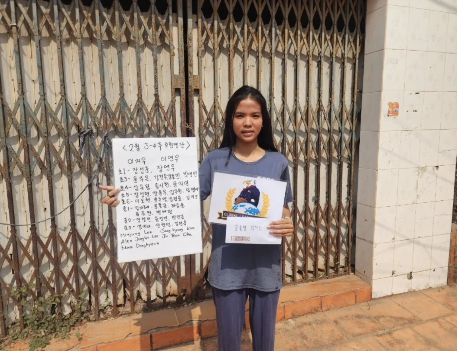 A young girl standing outdoors in front of a metal gate and holding a notebook with a cartoon graphic on the cover. She is pointing at a handwritten sign in Korean beside her.