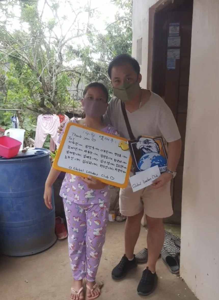 A man and a young girl wearing masks, holding handwritten notes, standing on a porch with greenery and laundry outside.