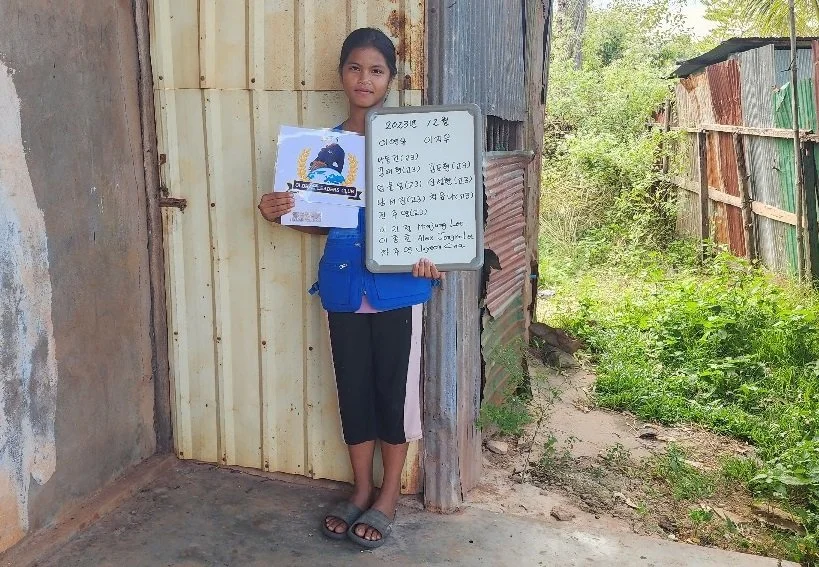 Young girl standing outside a wooden and corrugated metal shed, holding a whiteboard with handwritten text in a foreign language and a piece of paper with a colorful graphic, on a dirt path surrounded by greenery.