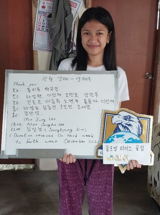 A young woman stands indoors holding a whiteboard with handwritten notes and a shield-shaped sign with a roaring eagle. She is smiling, wearing a white shirt and patterned purple pants.