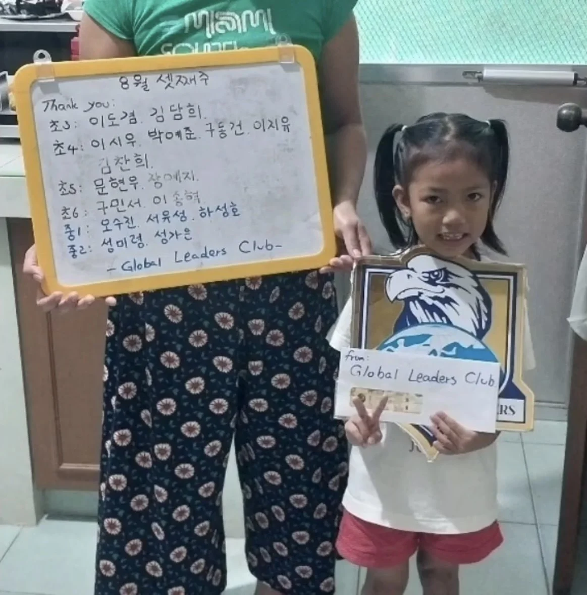 A young girl with pigtails holding a sign that says 'from Global Leaders Club', standing next to an adult holding a whiteboard with writing in Korean, both inside a room with a green and white background.