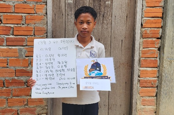A young boy standing outdoors in front of a wooden fence, holding a handwritten sign and a paper with a logo. The sign includes text written in Korean and English.
