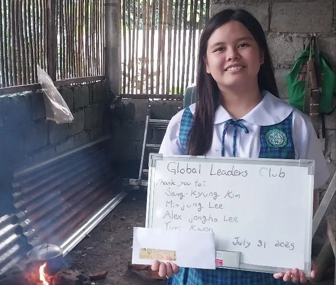 Young girl in school uniform holding a whiteboard with handwritten notes, standing inside a rustic room with bamboo and concrete walls.