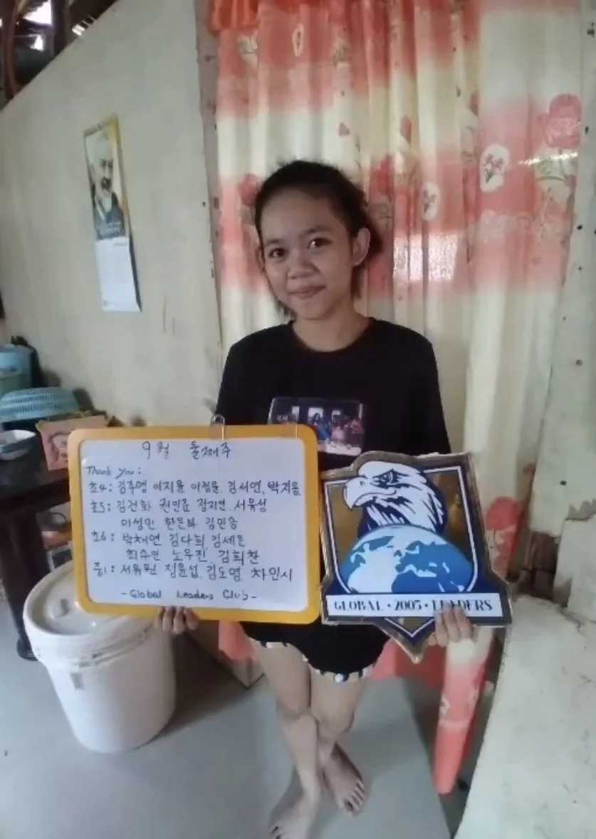 A young girl standing indoors, holding a whiteboard with handwritten text and a shield-shaped sign with an eagle and globe graphic. She is wearing a black shirt and shorts, with a pink curtain in the background.