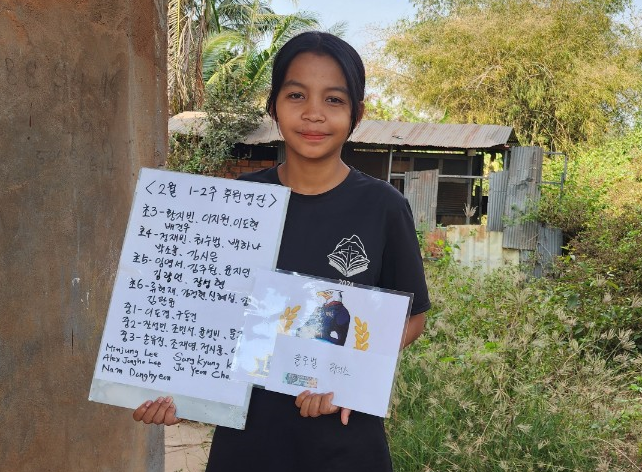 A young girl with black hair holding a paper with handwritten text and a card, standing outdoors near a rustic building and greenery.