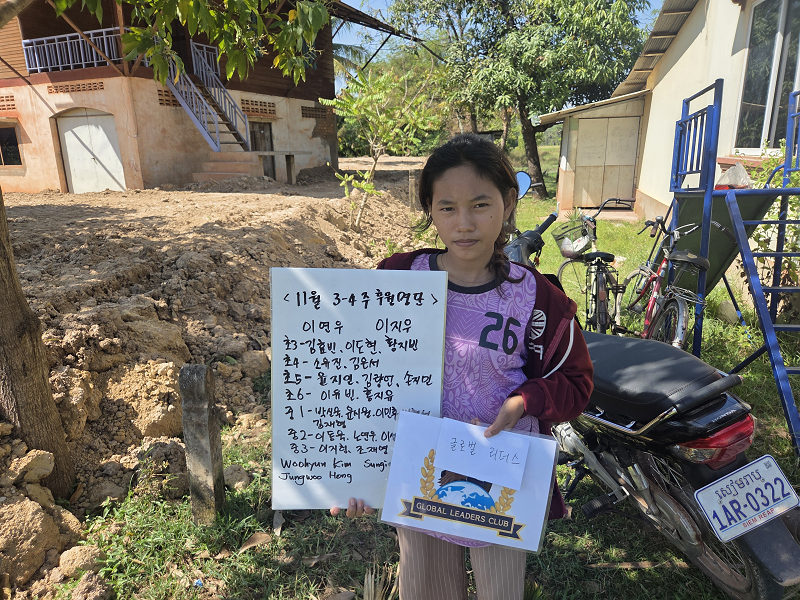 A young girl standing outdoors, holding a handwritten sign and a certificate. There are bicycles and a motorcycle behind her, a house and trees in the background.