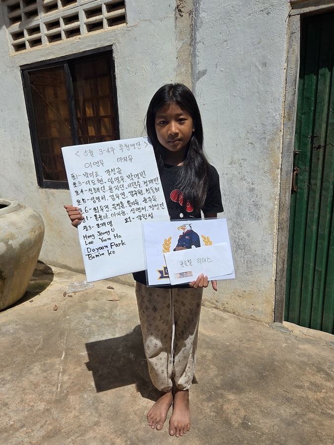 A young girl with long black hair standing outside on a concrete surface, holding a handwritten letter and a paper with a printed image of an eagle. She is barefoot and wears a black shirt with a red design and loose beige pants. The background inclu