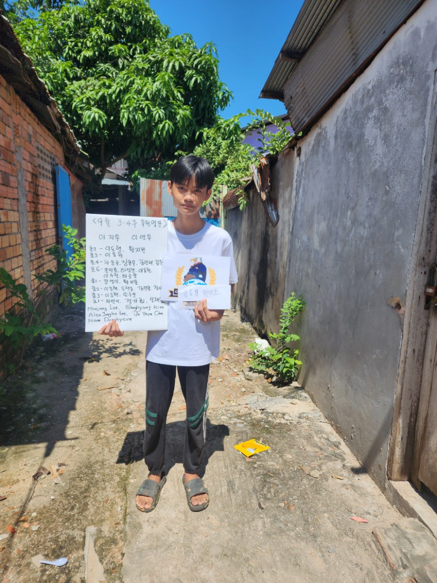 A young boy standing outdoors in a narrow alleyway, holding a handwritten sign in one hand and a smaller paper with text and an illustration in the other. He is wearing a white t-shirt, dark pants, and sandals. The alley is flanked by walls made of b