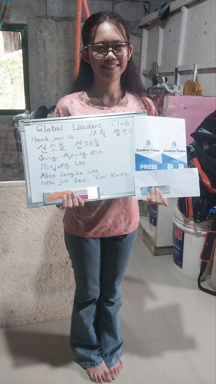 Young woman with glasses holding a whiteboard and envelope, smiling indoors, wearing a pink tie-dye shirt and jeans, standing barefoot on a tiled floor.
