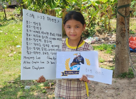 A young girl is holding a handwritten letter in Korean and a certificate or award with a globe and 'GLOBAL LEADERS' written on it. She is standing outdoors near trees and a garden.