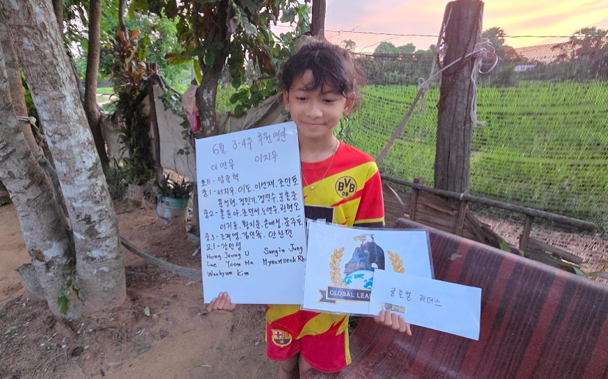 A young boy holding handwritten notes and a drawing, standing outside with trees and a field in the background during sunset.