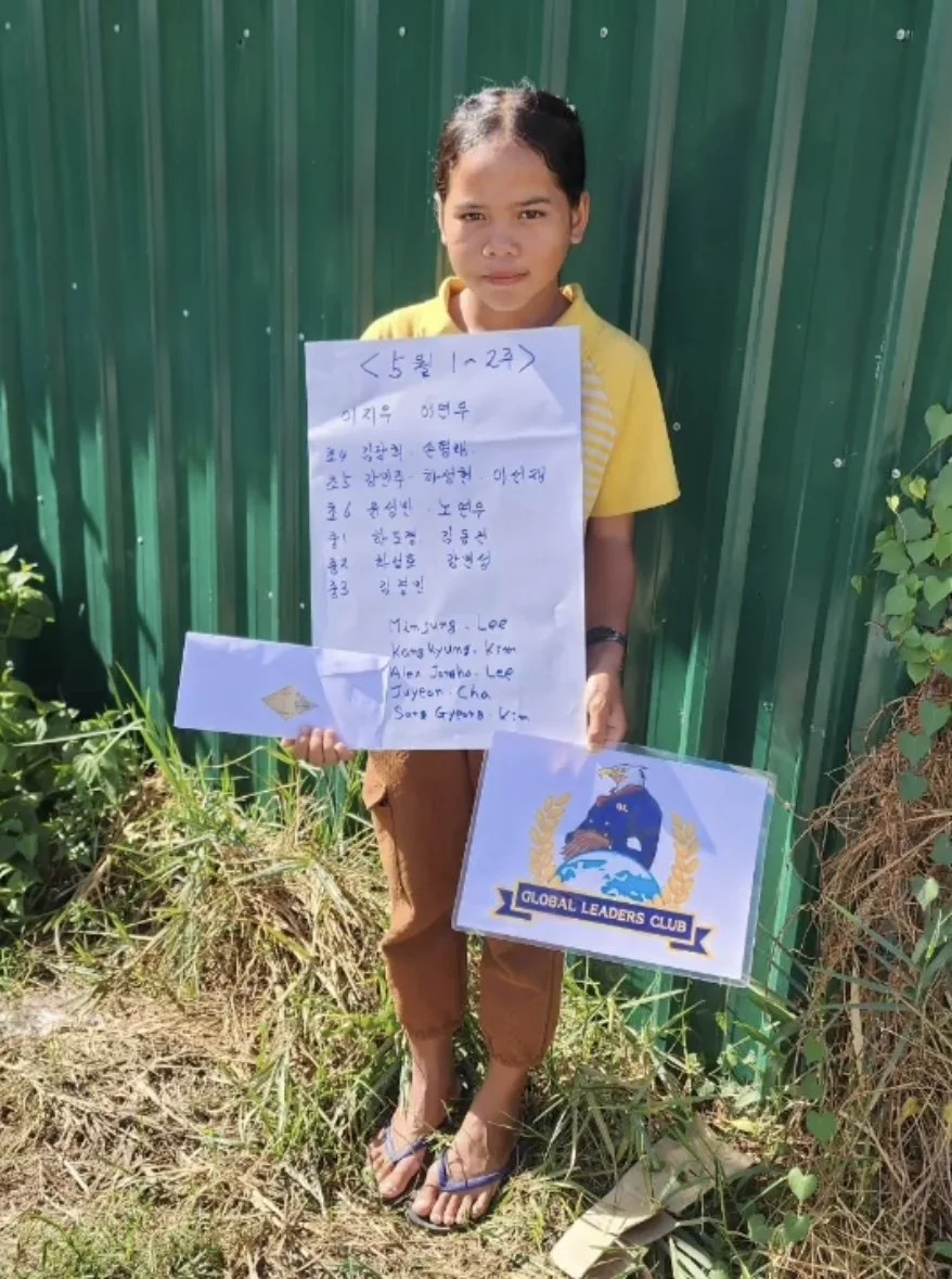 A young girl in a yellow shirt and brown pants holding a large handwritten note and a sign with the 'Global Leaders Club' logo, standing outdoors on grass in front of a green metal fence.