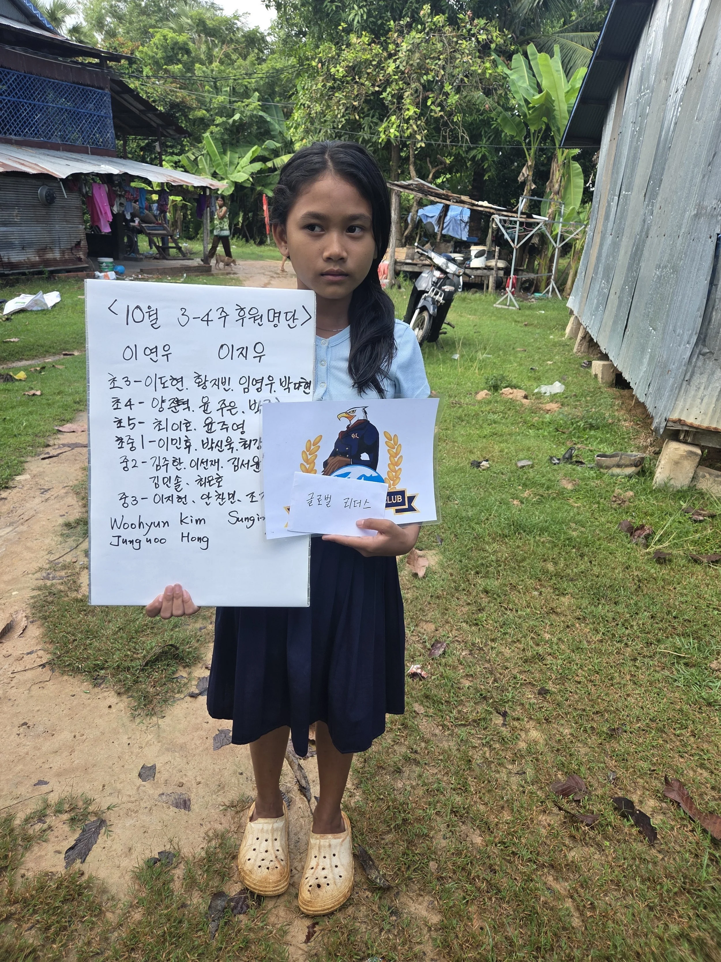 A young girl standing outdoors holding a white sign and a paper with a logo in front of a simple rural setting with houses, trees, and a motorcycle.
