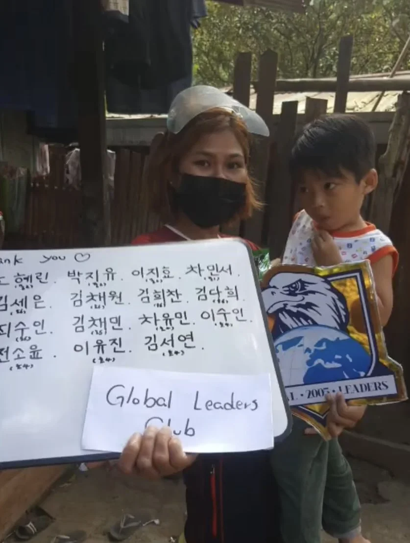 A woman wearing a black face mask and a clear plastic face shield holds a whiteboard with handwritten names and a note that says 'Global Leaders Club.' She stands next to a young boy in a sleeveless shirt who is holding a patch with an eagle and the 