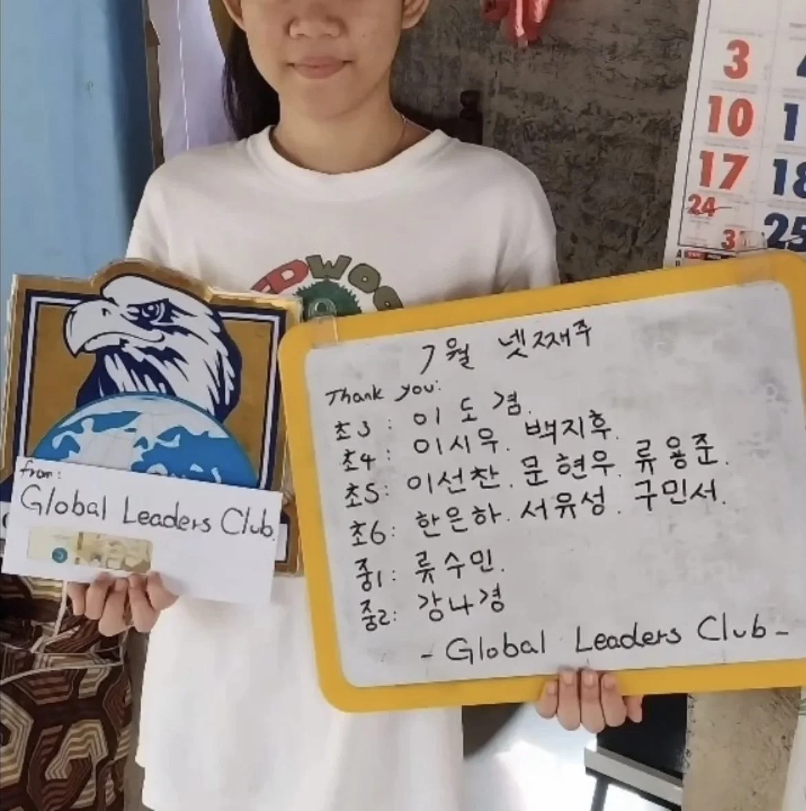 A young girl holding a sign that says 'Global Leaders Club' and a whiteboard with handwritten Korean text and a list of names, standing in a room with a calendar in the background.