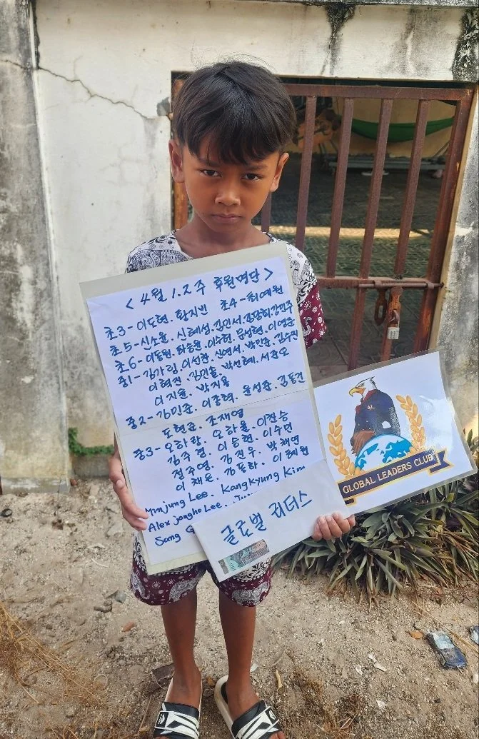 A young boy standing outdoors in front of a weathered white wall and a rusted gate, holding a handwritten letter in a foreign language and a sign with a logo for the Global Leaders Club, featuring an eagle and a globe.