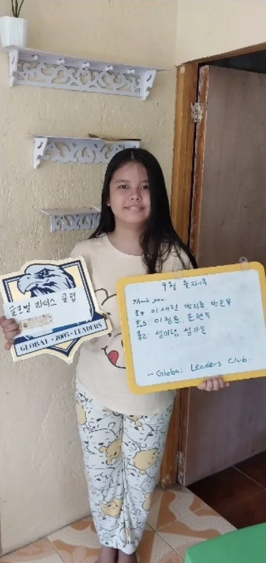 A young girl with long dark hair smiling, wearing pajamas with a teddy bear pattern, standing indoors near a beige wall with decorative white shelves. She is holding a plaque with an eagle logo and a note written on a yellowboard from the Global Lead