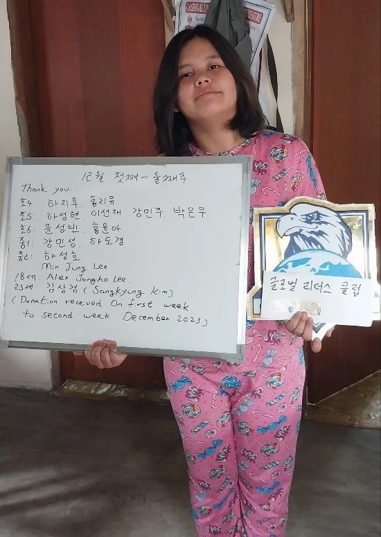 A young girl in pink pajamas with a colorful pattern holding a whiteboard with Korean writing and an image of an eagle, standing indoors with wooden walls and some papers hanging in the background.