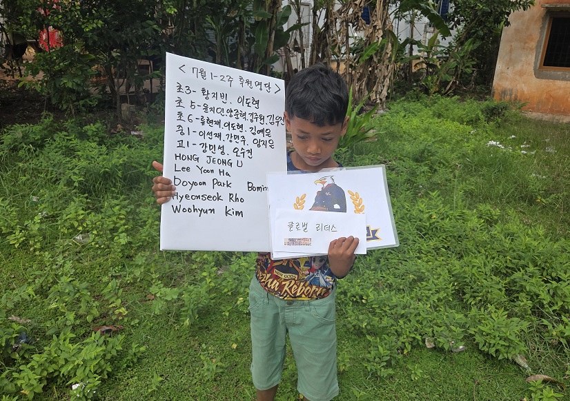 A young boy standing outdoors on a grassy area holding two papers with writing on them, surrounded by greenery and plants.