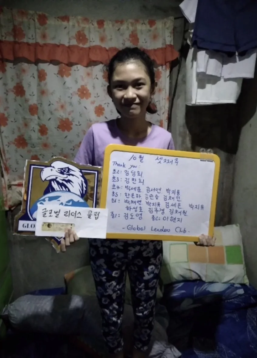 A young girl standing in a room holding a yellow whiteboard with handwritten notes and a sticker of an eagle. The room has floral curtains and hanging clothes, with a bed and pillow in the background.