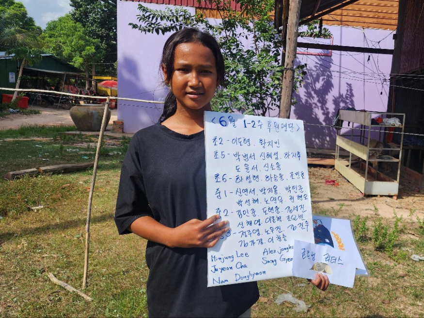 A young woman standing outdoors in a rural area, holding a handwritten sign in Hangul, the Korean alphabet. She is wearing a black T-shirt, and there is greenery and some simple structures behind her.