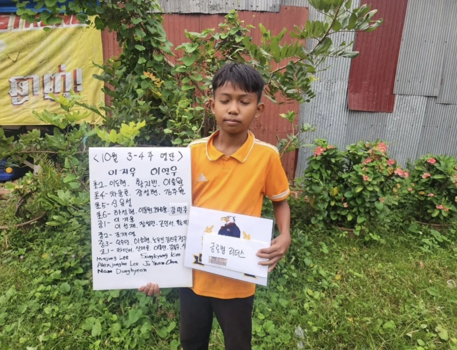 A young boy standing outdoors in a grassy area holding a whiteboard with handwritten text and a small booklet. He is wearing an orange shirt with white accents. Behind him are green plants, bushes with pink flowers, and a corrugated metal and wood fe