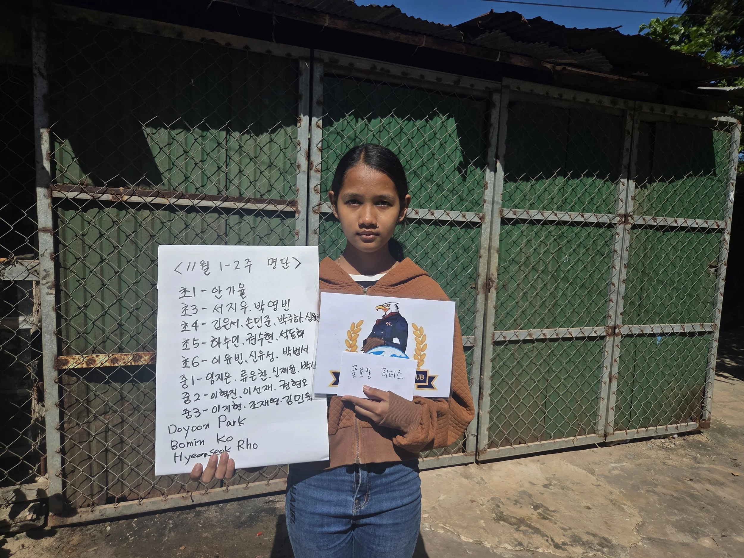 A girl standing outdoors in front of a rusted metal gate holding a white sign with handwritten text and a printed printout with a graphic of an eagle. She is wearing a brown hooded jacket and blue jeans.