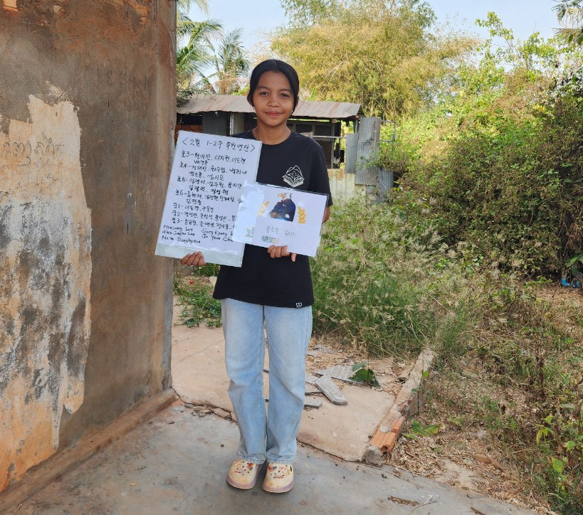A young girl standing outdoors, holding a handwritten letter and a drawing or card, in a rural area with trees and bushes in the background.