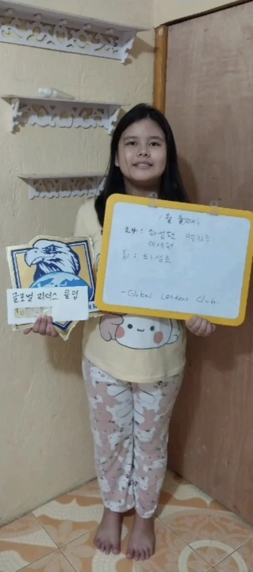 Young girl standing indoors, holding a yellow-framed whiteboard with handwritten notes in Korean and English, wearing pajamas with a cute character on the shirt, and pink pajama pants, standing barefoot on patterned tile floor.
