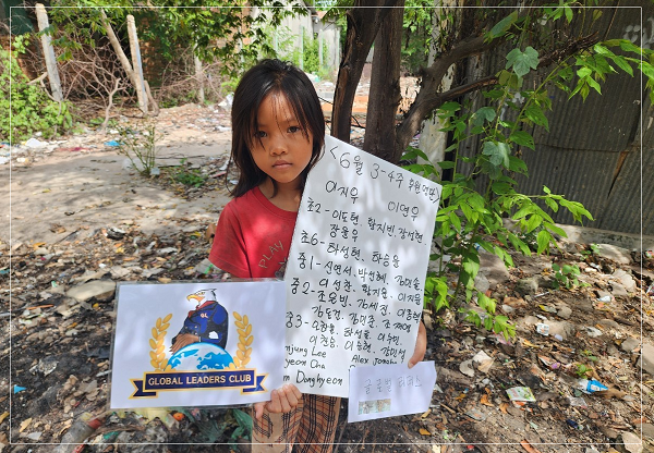 Young girl standing outdoors, holding a sign with Korean writing and a promotional card for the Global Leaders Club, surrounded by trees and dirt ground.
