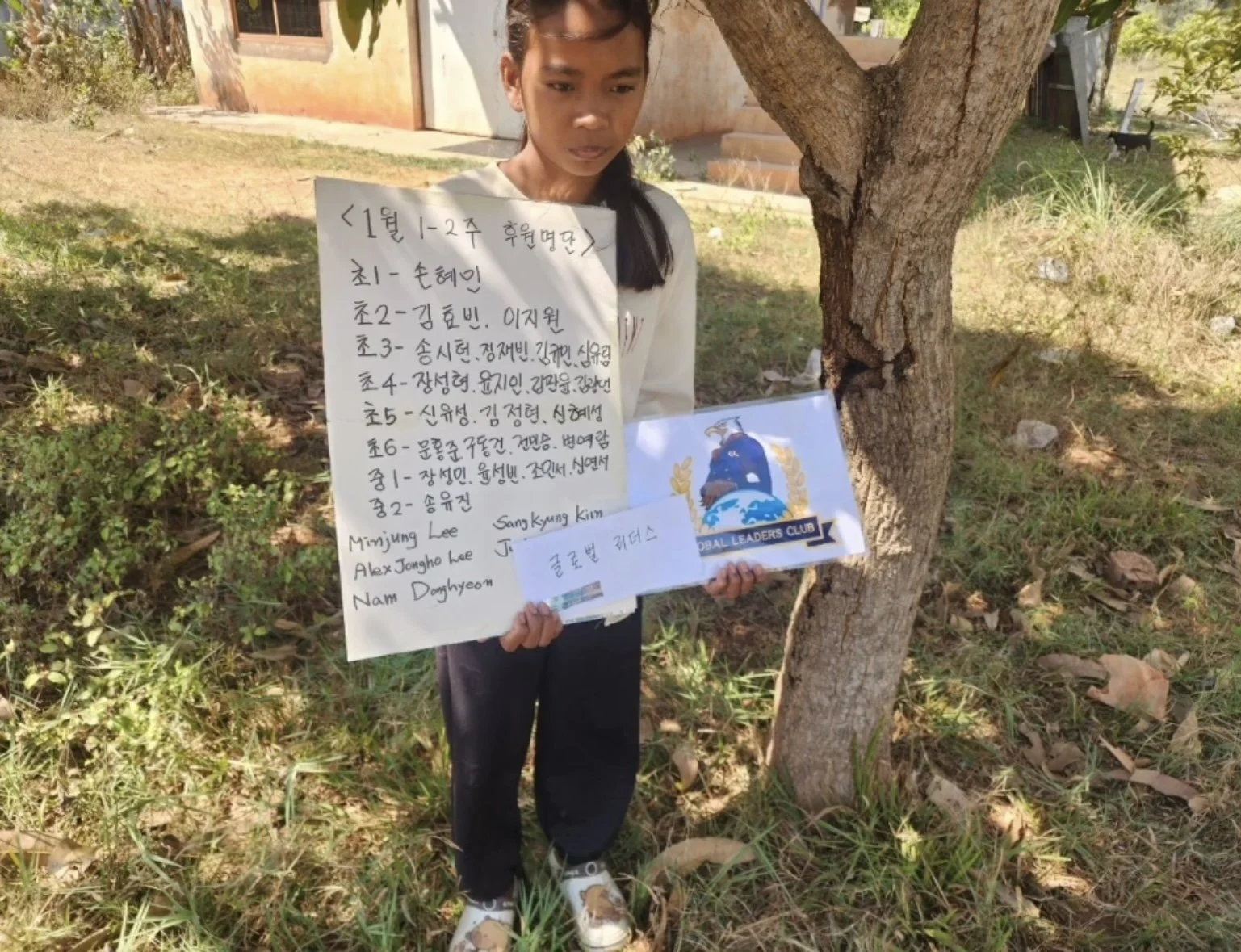 A young girl standing outdoors next to a tree, holding a sign with Korean writing and English names, with a logo for the Global Leaders Club on the sign. The background shows a grassy area, some rocks, and part of a building.