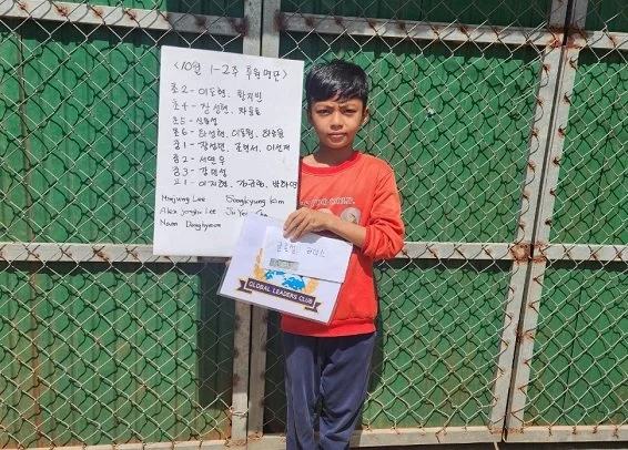 A young boy standing in front of a green chain-link fence, holding a sheet of paper with Korean writing and a colorful illustration, wearing a red long-sleeve shirt and dark pants.