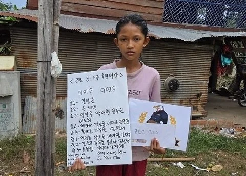 A young girl holding a sign with handwritten text and pictures, standing outdoors in front of a rustic wooden building.