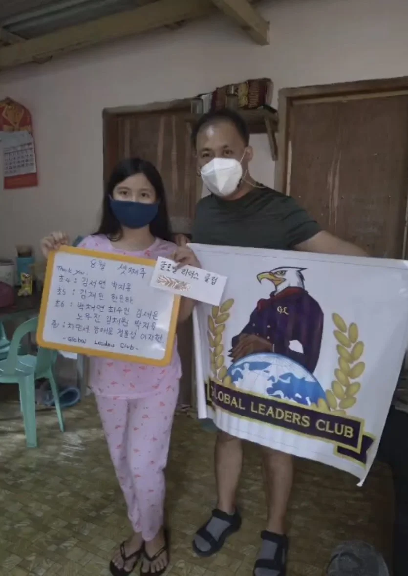 A girl and a man, both wearing masks, holding a sign with handwritten Korean text and a banner for the Global Leaders Club, inside a room with wooden walls and furniture.