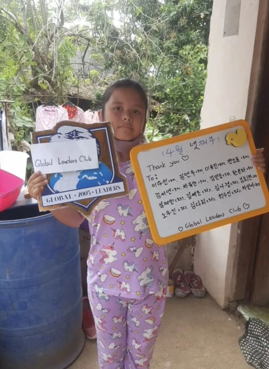A girl in pajamas holding a sign with a handwritten message and a shield labeled 'Global Leaders Club.'