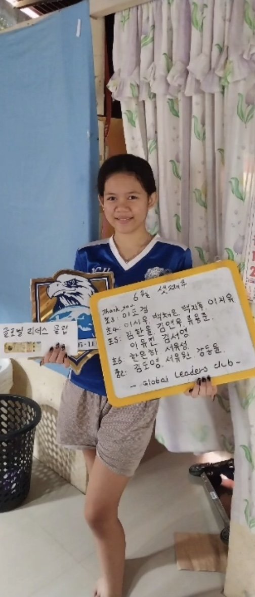 A young girl smiling and holding a whiteboard with handwritten message and a picture of an eagle. She is standing indoors near a curtain with green leaf patterns, with a black basket and laundry basket nearby.