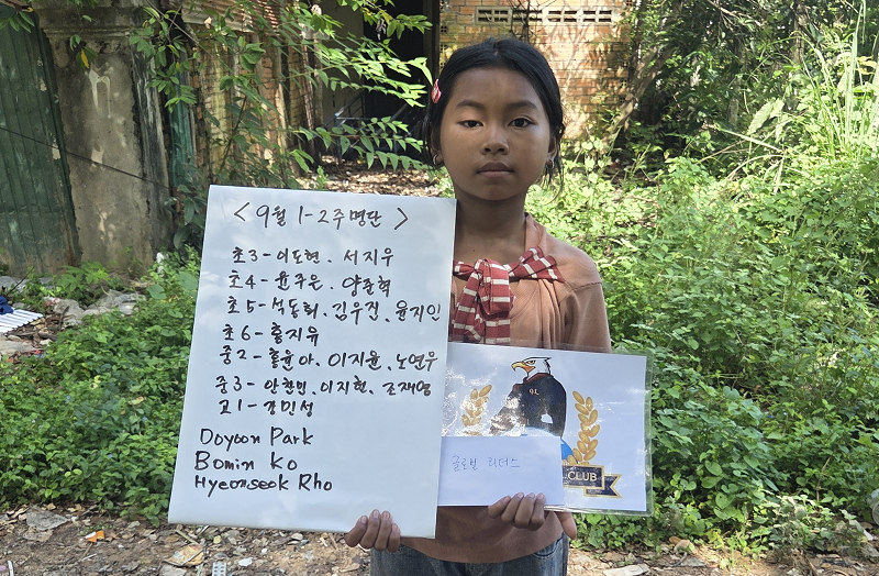 A young girl with dark hair and a pink bow in her hair, standing outdoors among green bushes, holding a handwritten note and a printed card. The note contains text in Korean and English, mentioning a schedule and locations such as Doyoon Park, Bomim 